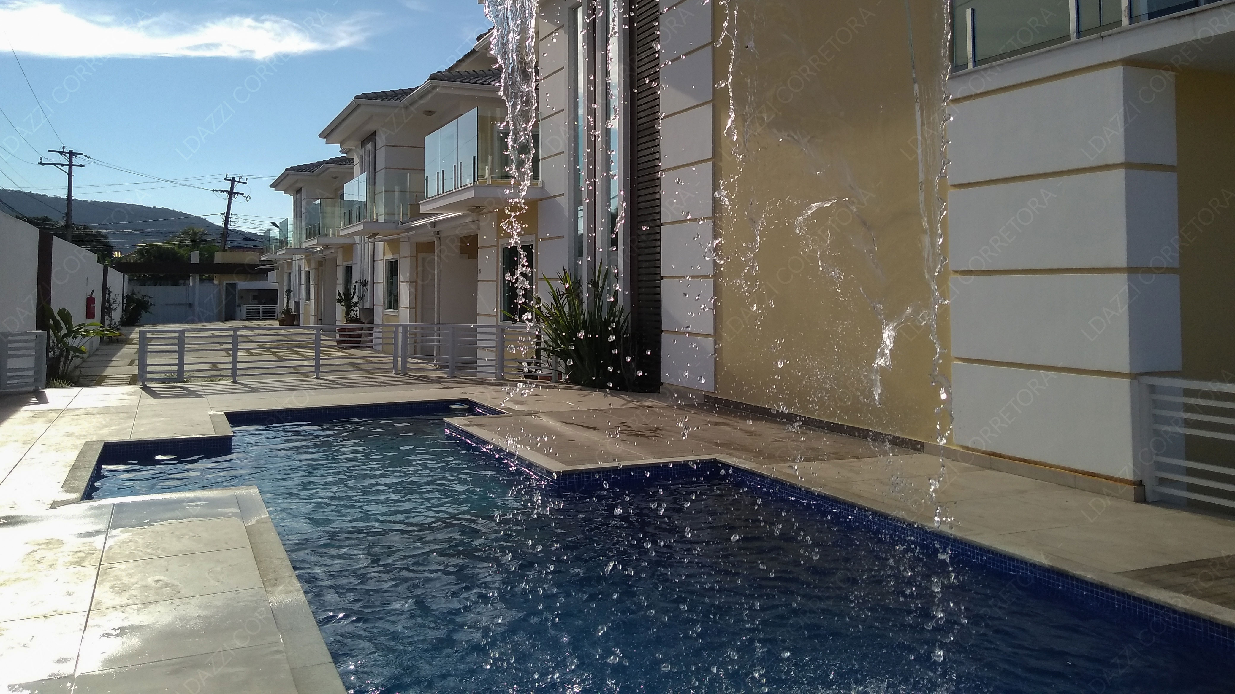 Piscina com cascata e fachadas do Condomínio Aurum Pescatore em Cabo Frio, sob luz natural suave